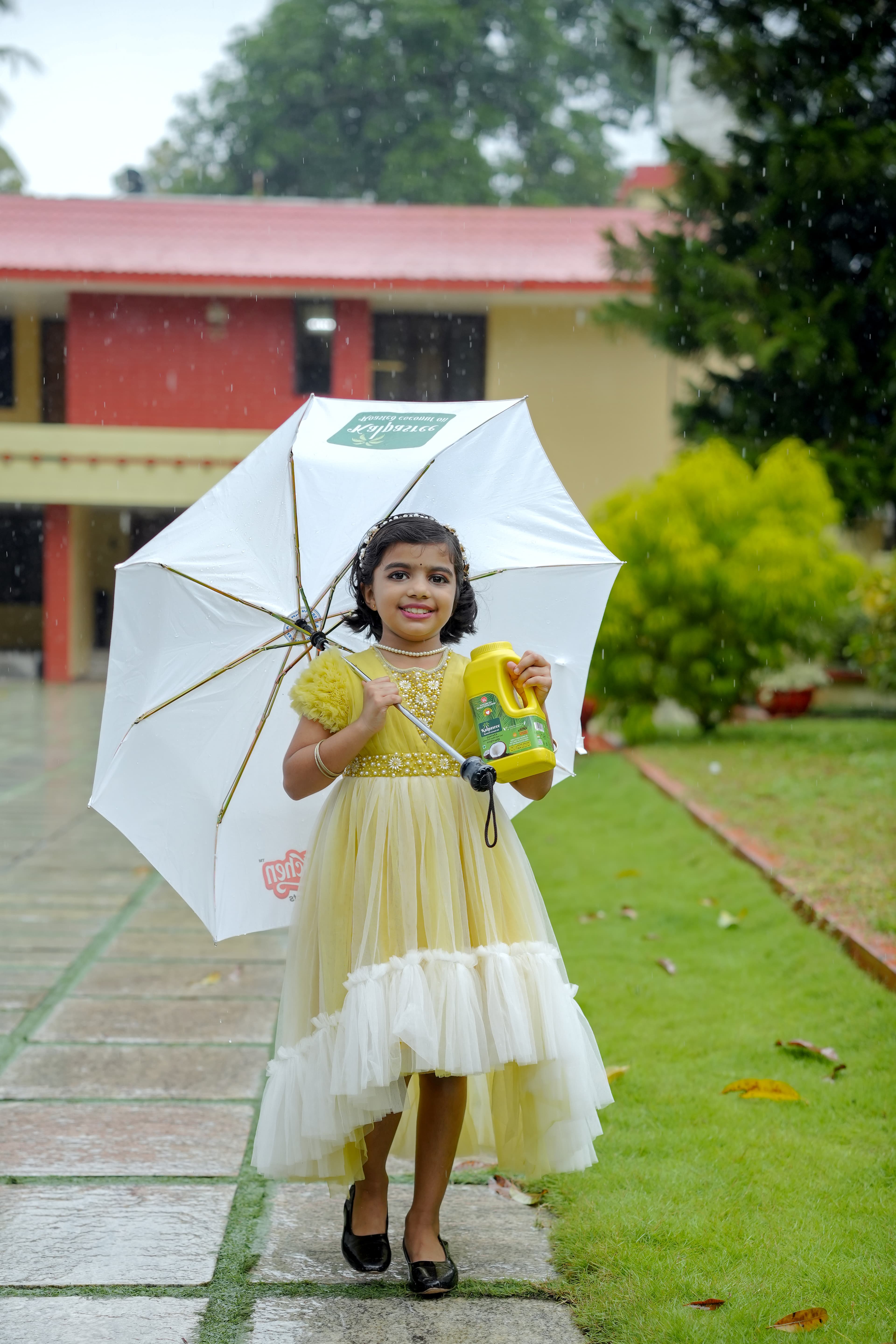 A child holding a Kalpasree can under a branded umbrella in the rain